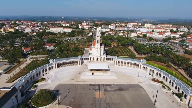 Majestic aerial footage flying away from the basilica of our lady of the rosary, revealing the expansive square and colonnade of the sanctuary of fatima in portugal on a sunny day