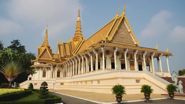 The Royal Palace in Phnom Penh Cambodia showcases ornate golden tiered roofs and white colonnades on a raised platform surrounded by manicured gardens and statues.