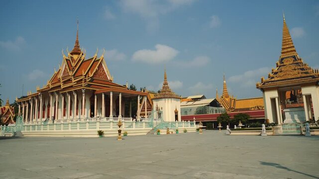 A wide daytime view of the Royal Palace complex in Phnom Penh, Cambodia, featuring ornate buildings with golden spires and a large paved plaza with few people.