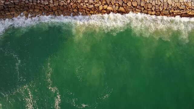 Stunning aerial top down view of turquoise ocean waves gently crashing against a long stone breakwater, creating white foam and a beautiful natural texture on a sunny summer day