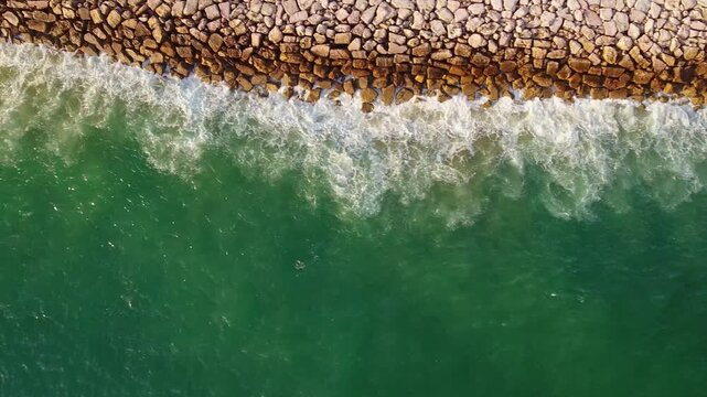 Stunning top down aerial perspective showing vibrant green ocean water with white foamy waves gently crashing against a long, rocky breakwater, creating a beautiful and hypnotic natural pattern