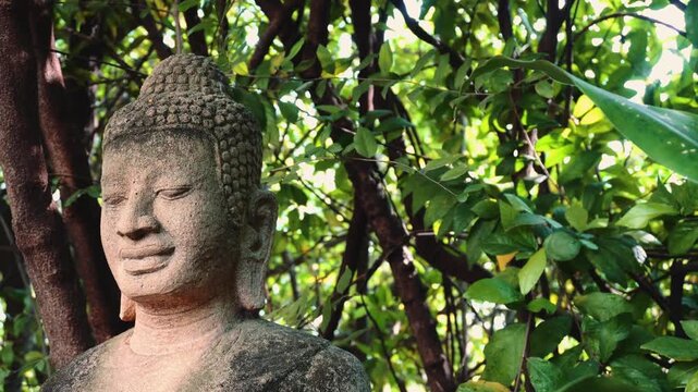 A large weathered stone Buddha statue with a serene expression stands outdoors amid dense green foliage in a garden setting in Phnom Penh, Cambodia during daytime.