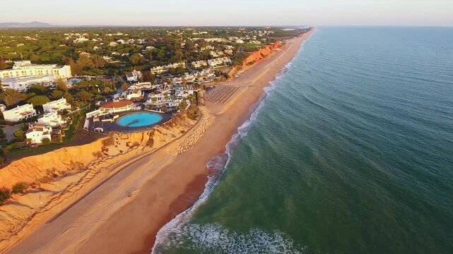 Majestic aerial view flying along the coastline of vale do lobo in the algarve region of southern portugal, showing beautiful sandy beaches, red cliffs, and luxury resorts at sunset