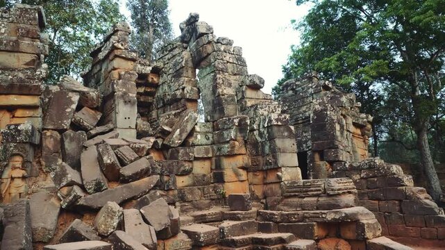 Extensive ancient stone temple ruins with weathered blocks and carved details surrounded by dense green foliage in a rural Cambodian archaeological site during daytime.
