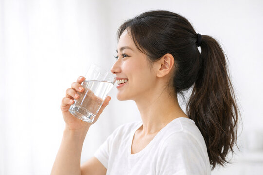 A woman in a sports bra drinking water from a clear glass, smiling and enjoying her refreshing beverage. She's wearing a white shirt and black pants, with natural lighting enhancing the vibrant scene.