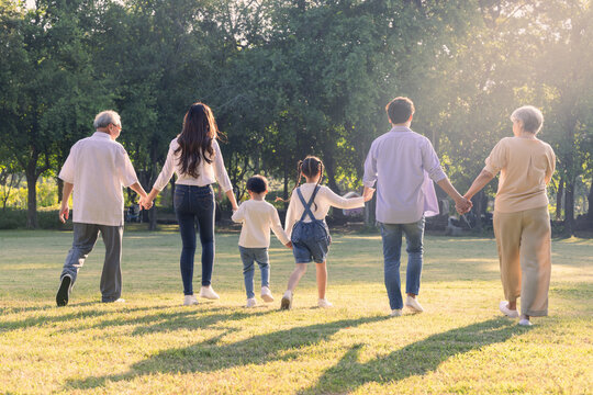 Multi-generational Asian family holding hands while walking through a sunny green park. Happy parents, children, and grandparents enjoying quality outdoor leisure time together during golden hour