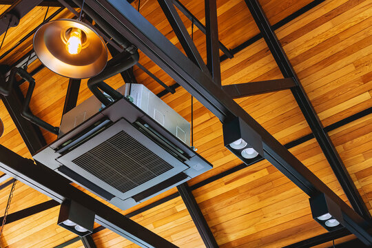 Modern cassette type air conditioner installed on loft ceiling with steel beams and wooden roof. Ceiling-mounted cassette air conditioning unit integrated into modern interior design. HVAC systems.