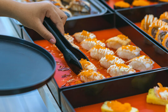 Woman customer choosing various sushi rolls with tongs at Japanese buffet restaurant. Hand of woman using tongs to pick fresh salmon nigiri sushi onto plate from food display tray at Japanese buffet.