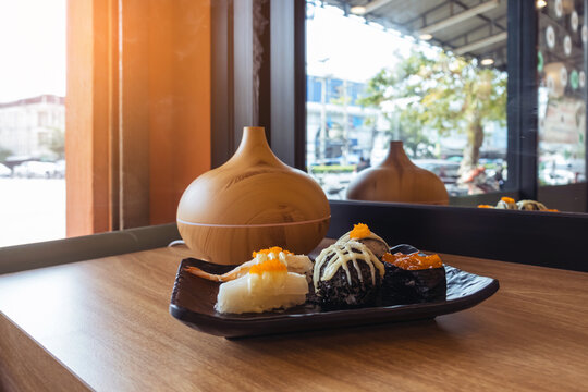 Sushi dish on window counter bar with ultrasonic humidifier. Assorted Japanese sushi, including nigiri and gunkan topped with roe next to wood grain aroma diffuser oil diffuser by window view.