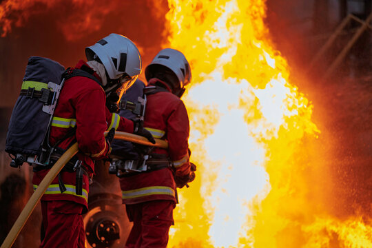 Professional firefighters in full protective gear bravely battle a massive, blazing inferno, holding a hose as intense orange flames and smoke consume the background in an emergency.