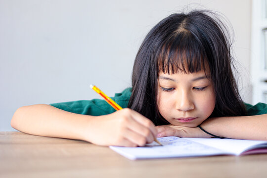 Focused young Asian girl studying at home, resting her arm on a wooden desk while diligently writing in a notebook with a pencil. Primary school student concentrating on homework.