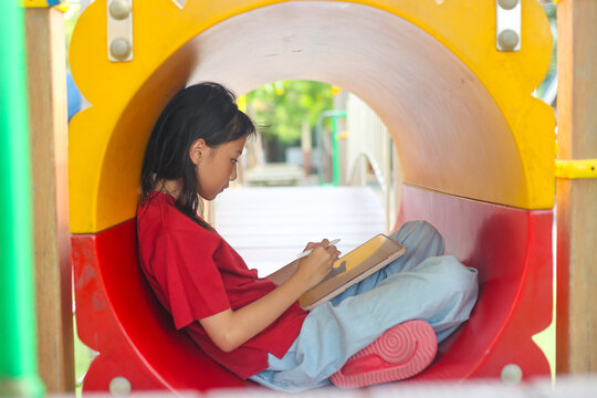Selective focus vibrant outdoor shot captures a focused Asian kid sitting inside a colorful playground tunnel. The girl in a red shirt is deeply engaged drawing on a digital tablet with a stylus.