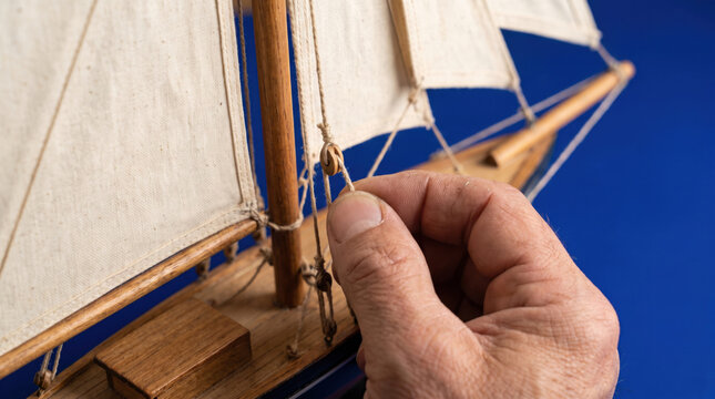 Hand model boat rigging sail wooden nautical miniature craft rope canvas close view of adult hand adjusting rigging on wooden model sailboat with calm blue background and detailed handmade texture