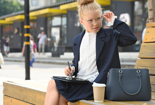 A little girl dressed as a businesswoman sits on a wooden bench outdoors