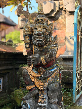 A stone statue of a fierce guardian figure, adorned with intricate carvings and colorful embellishments. The statue is set against a backdrop of lush greenery and traditional architecture.