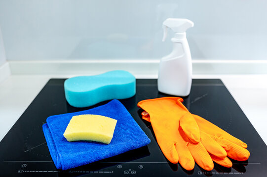 Cleaning tool products on countertop for home hygiene maintenance. Orange rubber gloves, blue microfiber cloth, sponge and detergent spray bottle on induction cooktop in modern kitchen