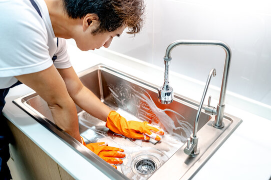 Asian male housekeeper with orange rubber glove scrubbing on kitchen sink surface with soap suds spreading around. Cleaning stainless steel sink for hygiene. Housework chores in the kitchen