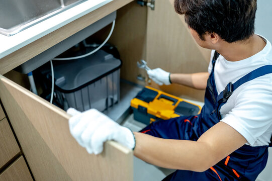 Male plumber with work tools opening wooden cabinet checking grease trap box and pipe connection under the kitchen sink. Handyman prepares to fix the plumbing system.