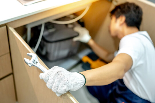 Asian male plumber with glove and adjustable wrench in his hand opening wooden cabinet checking grease trap box and pipe system under the kitchen sink. Handyman fixing plumbing system in the kitchen.