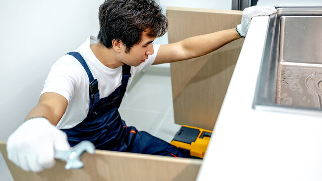 Asian male plumber with glove and adjustable wrench in his hand opening wooden cabinet checking pipe system under the stainless steel sink. Handyman fixing plumbing system in the kitchen.
