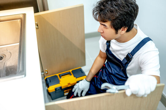 Asian male plumber with glove and adjustable wrench in his hand opening wooden cabinet checking pipe system under the stainless steel sink. Handyman fixing plumbing system in the kitchen.