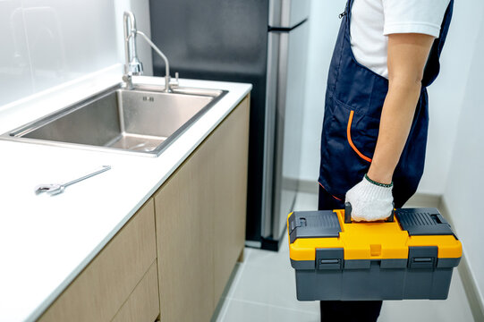 Male plumber holding yellow toolbox in the kitchen. Handyman in uniform prepares to fix plumbing system under stainless steel sink in wooden cabinet. Home maintenance service