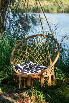 A woven hanging chair with a patterned cushion overlooks a calm river amid lush greenery. Ideal for relaxation, outdoor living, garden design and wellness concepts.