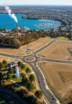 Aerial View of Coastal Development with a Lighthouse, Roundabout, Marina, and Industrial Chimney in a Sunny Landscape