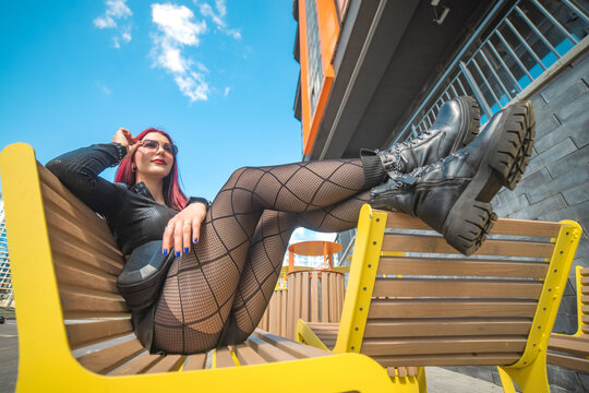 Edgy red haired girl in fishnet tights relaxing on yellow bench with sky copy space