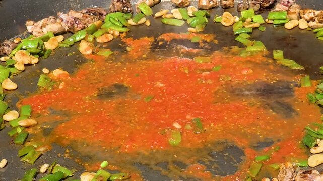 Close-up shot of simmering tomato puree sofrito with green flat beans and lima beans in a large flat pan during a traditional Spanish paella cooking competition outdoors.