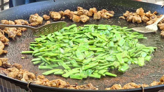 Close-up shot of stirring green flat beans in oil at the center of a traditional paella pan while fried chicken pieces sit along the rim during Spanish paella cooking process.