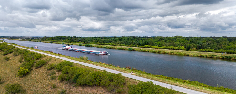 Cargo barge travels along Mittellandkanal through green landscape beneath dramatic summer clouds. Inland waterway supports freight transport, navigation and logistics across northern Germany