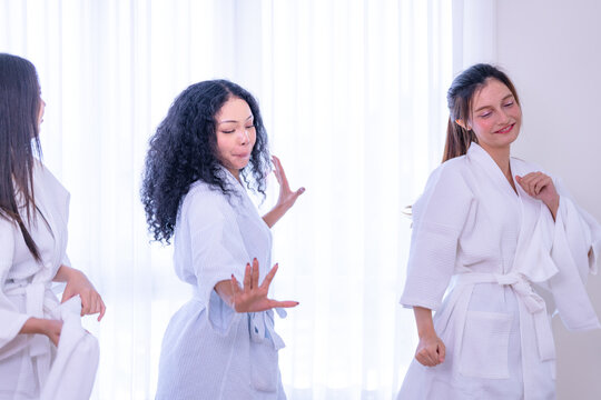 Full length shot of three young multiethnic women in white bathrobes messing about together while the Asian friend backs away giggling with her palms out in a bright white spa room.