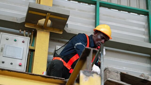 African industrial worker wearing safety helmet and vest talks on walkie talkie near control panel box inside factory. Engineer or technician coordinates manufacturing operation