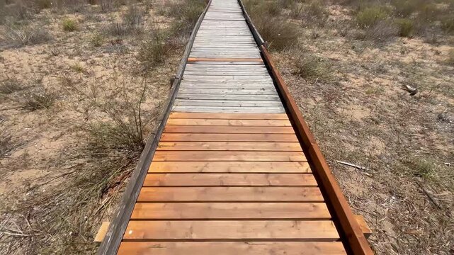 Wooden boardwalk leading through the sandy landscape of Foce Belice Nature Reserve in Sicily, Italy, surrounded by Mediterranean vegetation on a sunny summer day.