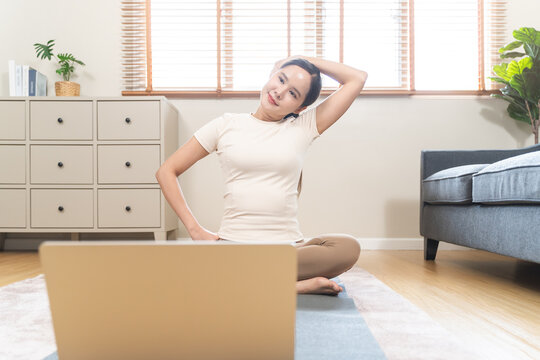 Happy asian young woman practicing yoga stretching neck exercise on mat watching online tutorial on laptop computer at home living room for office syndrome relief and wellness.