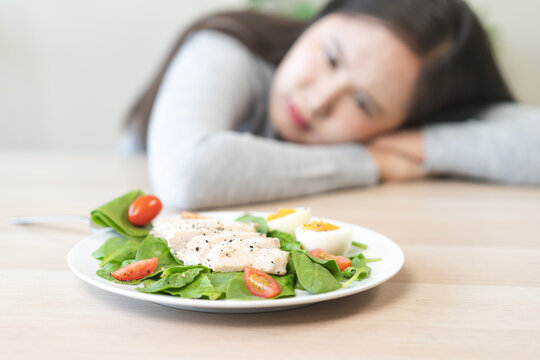 Sad and bored young Asian woman leaning on a wooden table at home, looking unhappy with her healthy salad plate, concept of diet stress, eating disorder, and weight loss struggle.