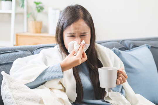 Sick concept, tired asian young woman have a fever, flu and hand in use tissues paper sneezing nose, runny and holding hot mug  while sitting on sofa at home. Health care with disease, pandemic virus.