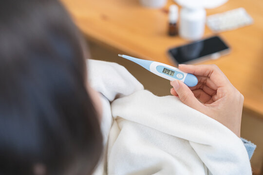 Close-up of sick Asian woman hand holding a digital thermometer showing high body temperature of 39.7 degrees celsius, female wrapping in white blanket having a severe fever at home
