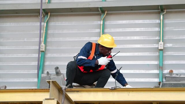 Industrial worker in a yellow hardhat and safety vest crouches on a steel beam to coordinate facility maintenance using a walkie-talkie at a warehouse terminal