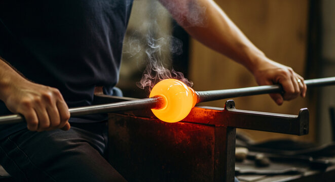 A skilled glassblower shaping a glowing molten glass sphere on a blowpipe in a workshop.