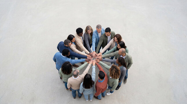 A diverse group of people standing in a circle, stacking their hands together in a gesture of unity and teamwork, viewed from above.