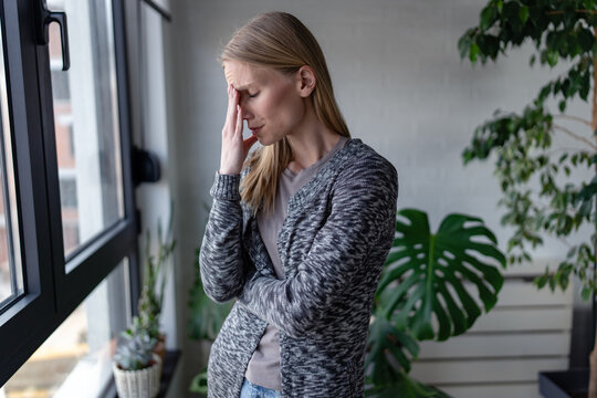 Exhausted woman covering her face while taking a break near a window. Burnout, work stress, emotional exhaustion, fatigue, pressure and mental wellbeing concept