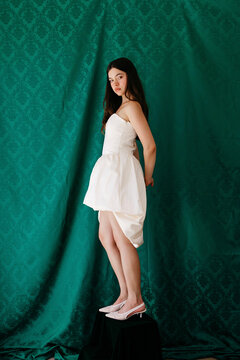 Young woman posing in white strapless dress against damask background