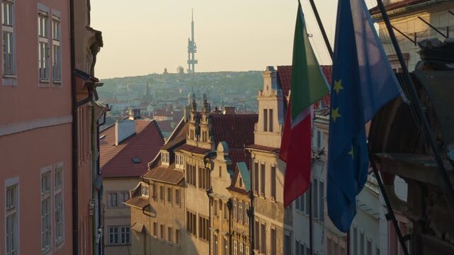 European Union flag hangs on a historic Prague street with traditional architecture. The city skyline features a famous television tower in morning light. Ideal for Czech travel and tourism.