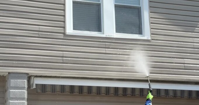 A person power washes beige vinyl siding, sweeping a strong water stream across the surface to remove dirt and grime. 