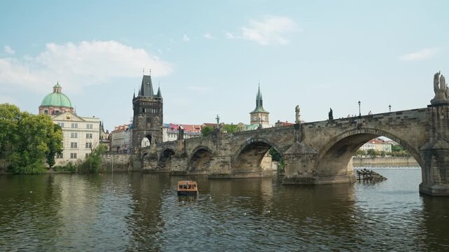 Historic Charles Bridge stands over Vltava River in Prague. A small tourist excursion boat sails under stone arches past city landmarks. Ideal for European vacation and tourism travel design.