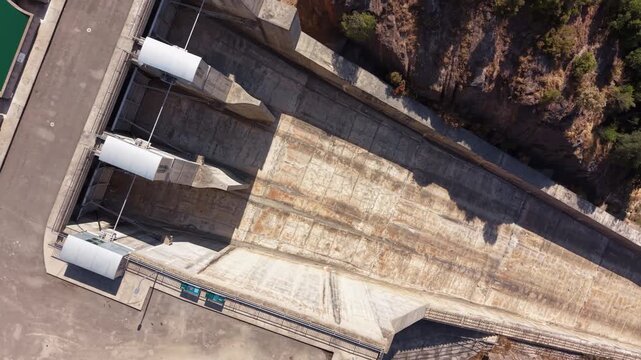Overhead view of Baells Dam spillway gate structures at the crest, showing concrete texture, mechanical infrastructure, green reservoir water, and rocky gorge in Catalonia, Spain