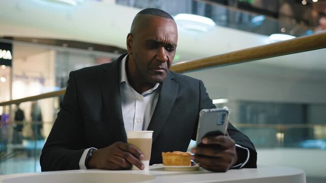 African American middle-aged business man pensive looking in mobile phone app drink coffee breakfast dinner with pie in cafe businessman male serious reading news in smartphone drink tea in cafeteria