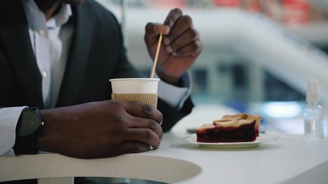 Close up male hands holding cup of hot coffee stirring sugar in mug unrecognizable African American busines sman visitor client stir drink cappuccino drinking with berry pie tasty cake at cafe table